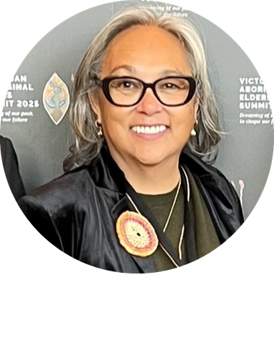 Smiling woman with gray hair and black-framed glasses, wearing a black jacket and circular badge, in front of a backdrop.